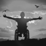 Guy in wheelchair extending his arms to the horizon with birds flying in the background